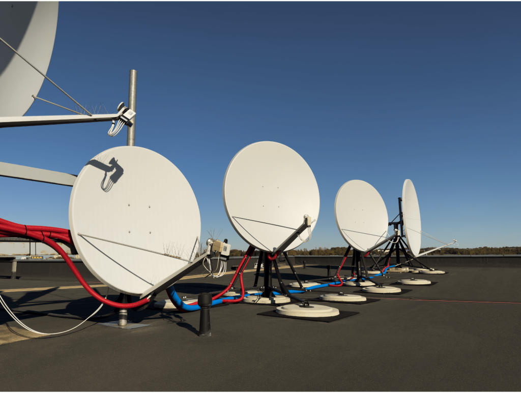 Row of rooftop satellite dishes under a clear blue sky, connected with red and blue cabling, used for broadcast signal transmission and telecommunications infrastructure.