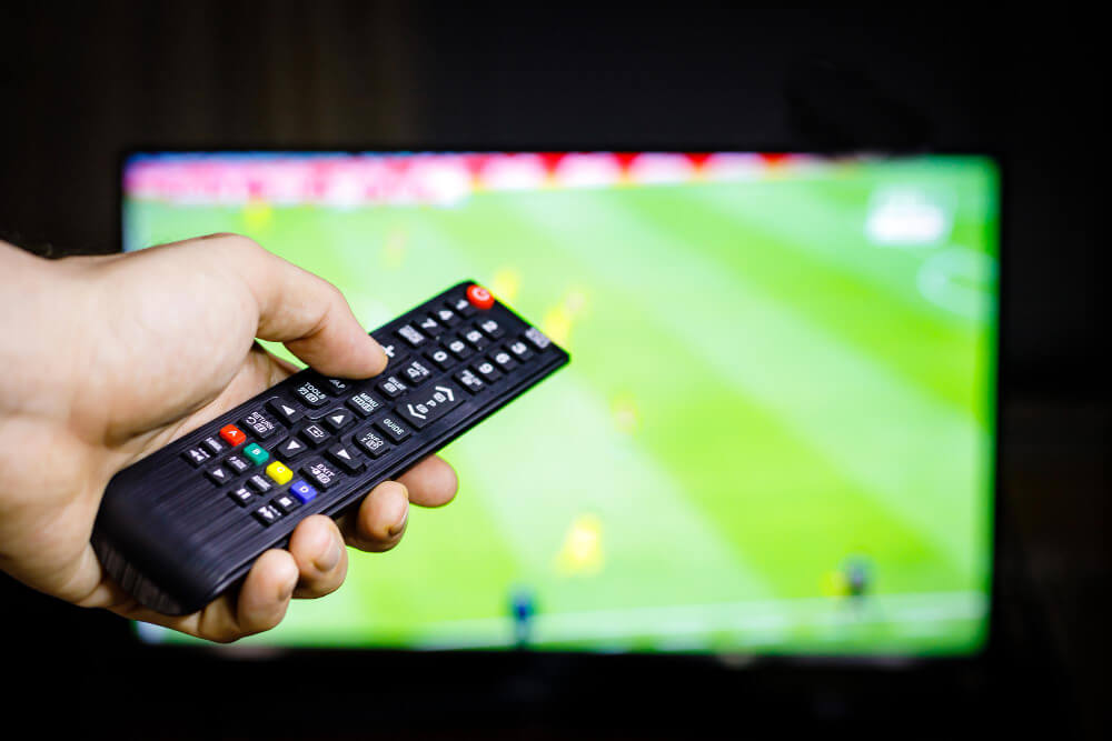 Hand holding a TV remote control pointed at a television screen displaying a soccer match in the background.